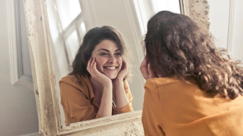 Woman with curly brown hair smiling at her reflection in the mirror, wearing an orange top, promoting self-confidence and wellbeing.