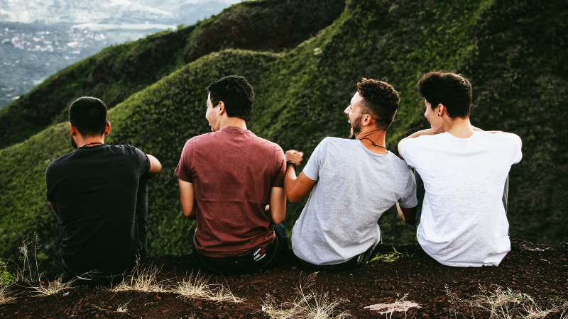 Back view of four men sitting together laughing and looking out over a green mountain landscape, representing male friendship, connection, and wellbeing.