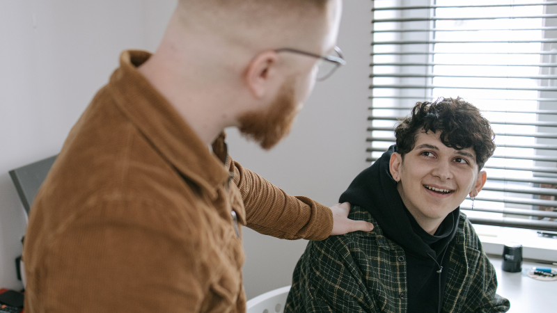 One man placing a supportive hand on another man’s shoulder while talking at an office desk, symbolising counselling, guidance, and emotional support.
