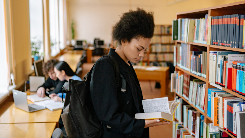 Female student reading a book in a university library, with two other students studying in the background, illustrating focus, learning, and academic wellbeing.