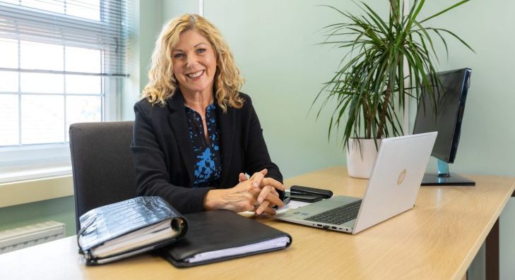 Bridget Walford, owner of The Eaves Counselling and Psychology, smiling at her desk with a laptop and house plant, representing professional leadership and approachability