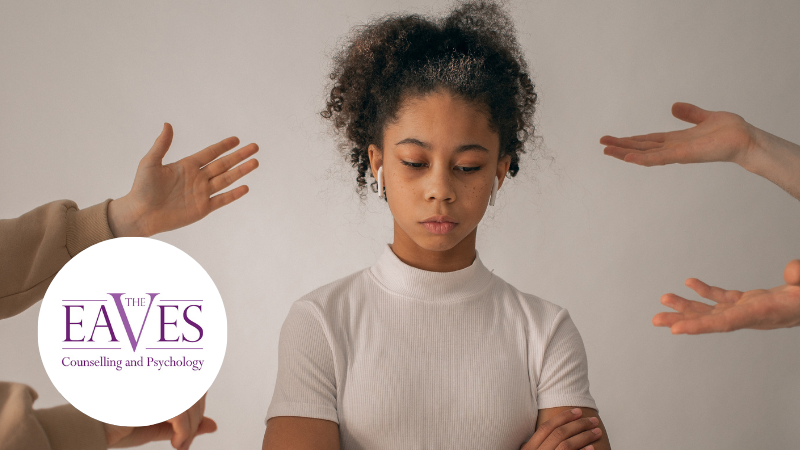 Anti-Bullying Week One Kind Word image showing a distressed teenage girl wearing headphones with her arms crossed while pointing hands surround her, representing bullying and emotional isolation