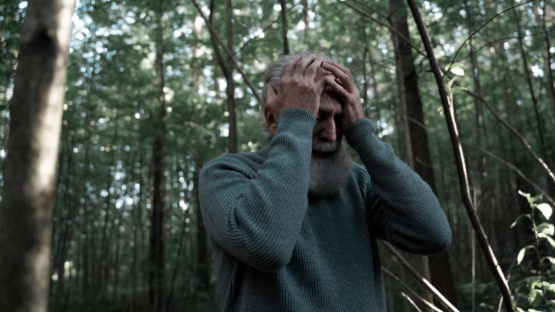 Man with a beard holding his head in his hands in a forest, experiencing a panic attack