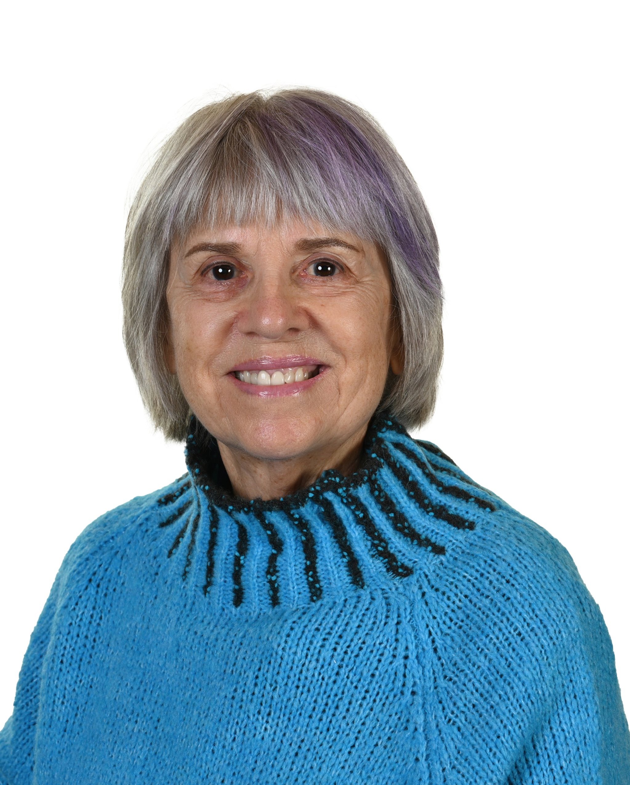 Anne Head, with short, gray and purple hair is wearing a blue knit sweater featuring a black and blue striped collar, set against a plain white background.