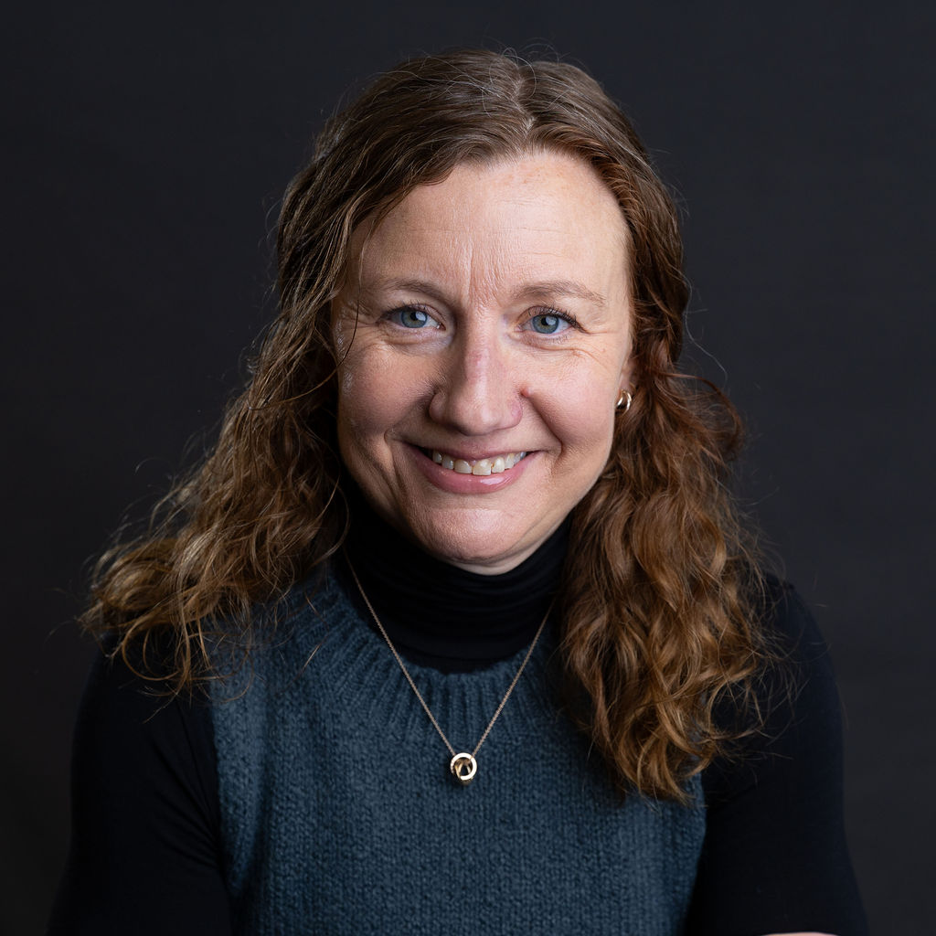 A woman with wavy brown hair and blue eyes, smiling at the camera, exudes Charlie Fancy charm. She wears a dark sleeveless top over a long-sleeve black shirt, highlighted by a gold necklace against the plain dark background.