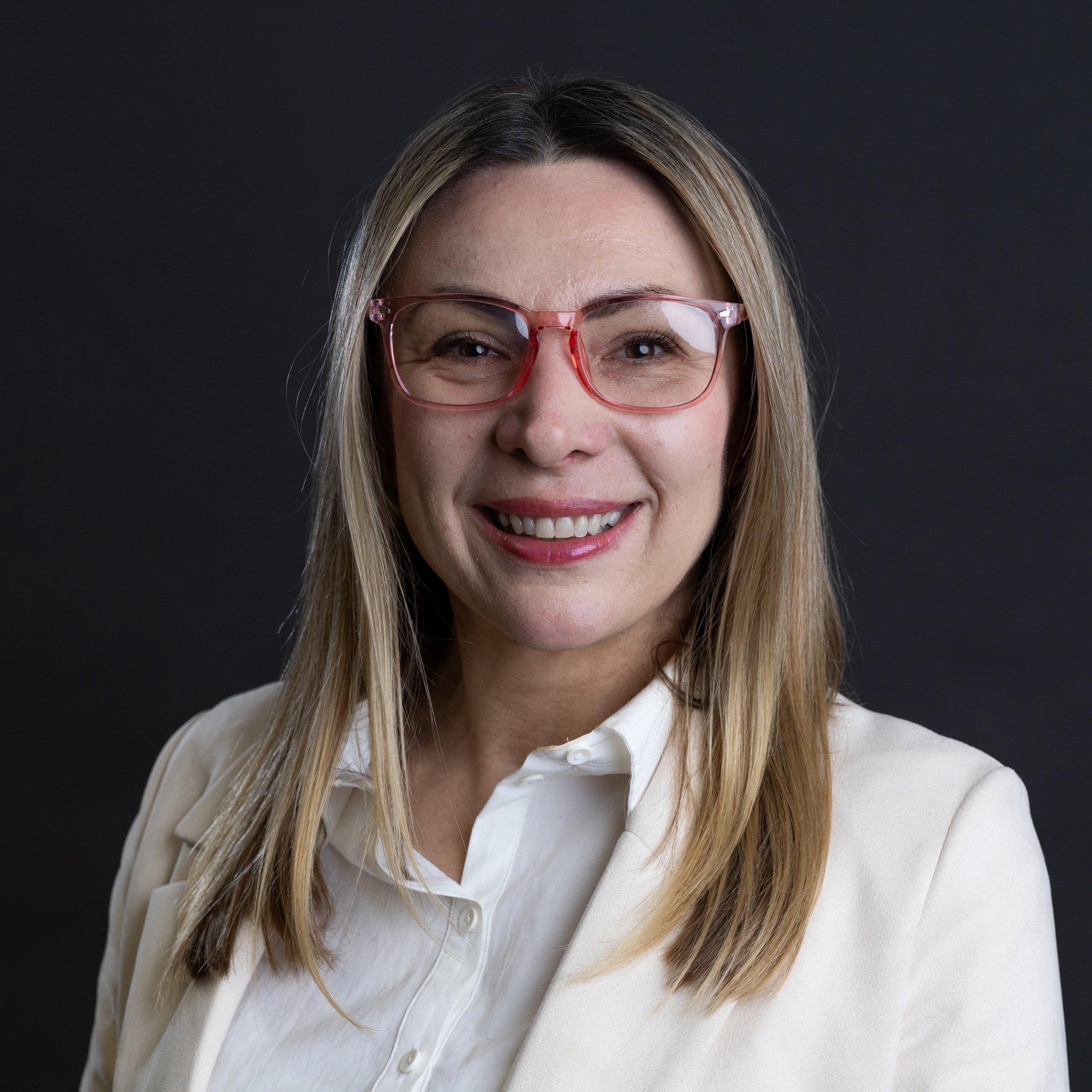 Marzena Steels, a woman with long blonde hair, smiles at the camera in her white blouse and jacket. She stands against a dark background, her red-framed glasses adding a pop of colour to the scene.