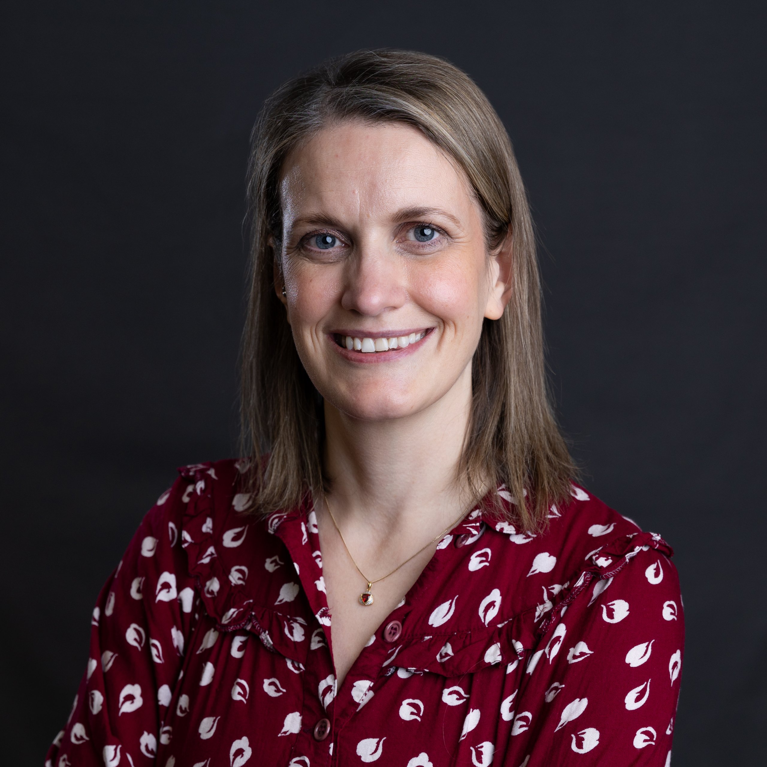 Dr. Melanie Orchard, a woman with shoulder-length hair, smiles at the camera. She wears a maroon blouse adorned with a white pattern and wears a small necklace against the dark gray background.