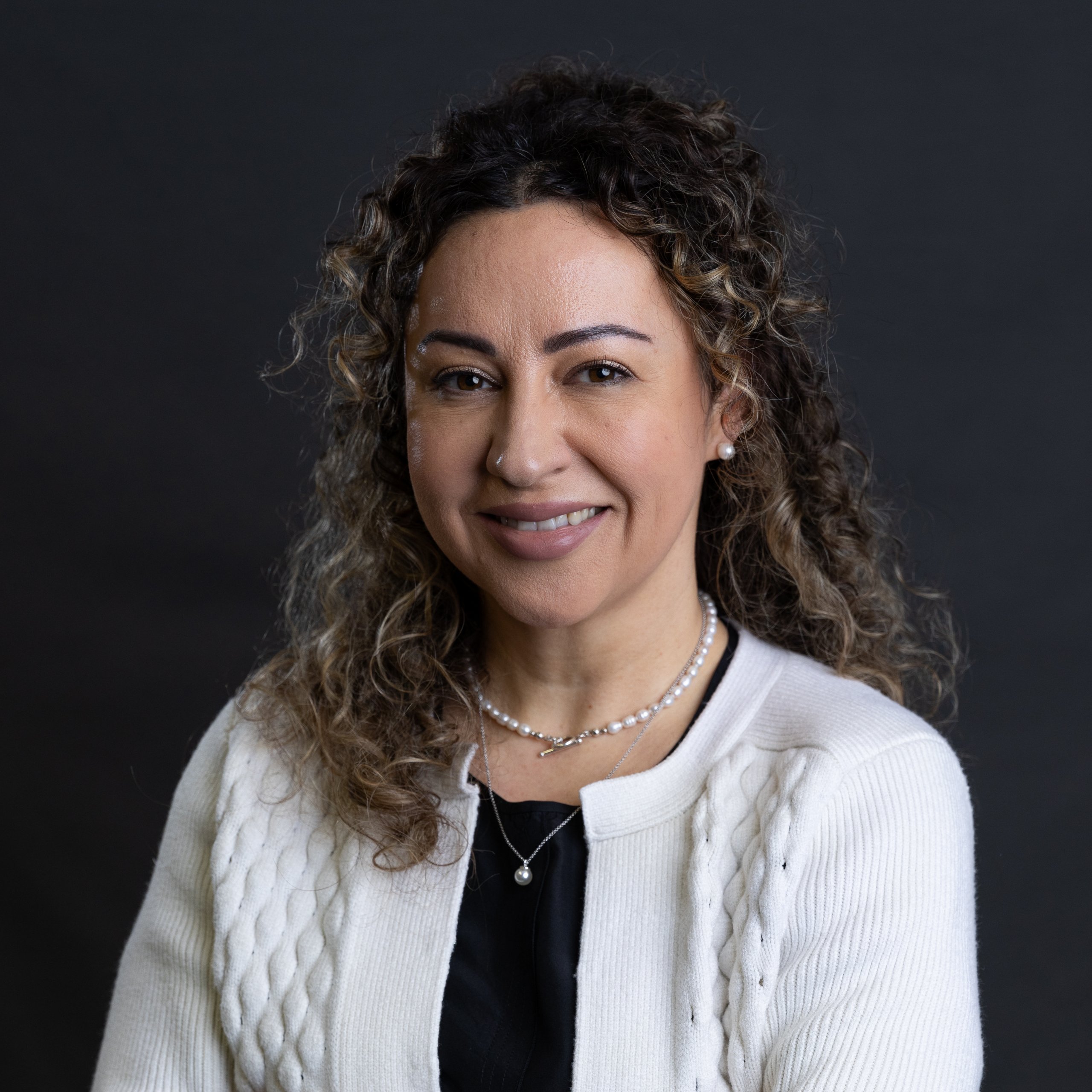 Dr. Simona Chereji, with curly hair, smiles warmly while wearing a white cardigan over a black top, elegantly accessorized with a pearl necklace. The plain dark background accentuates her poised presence.