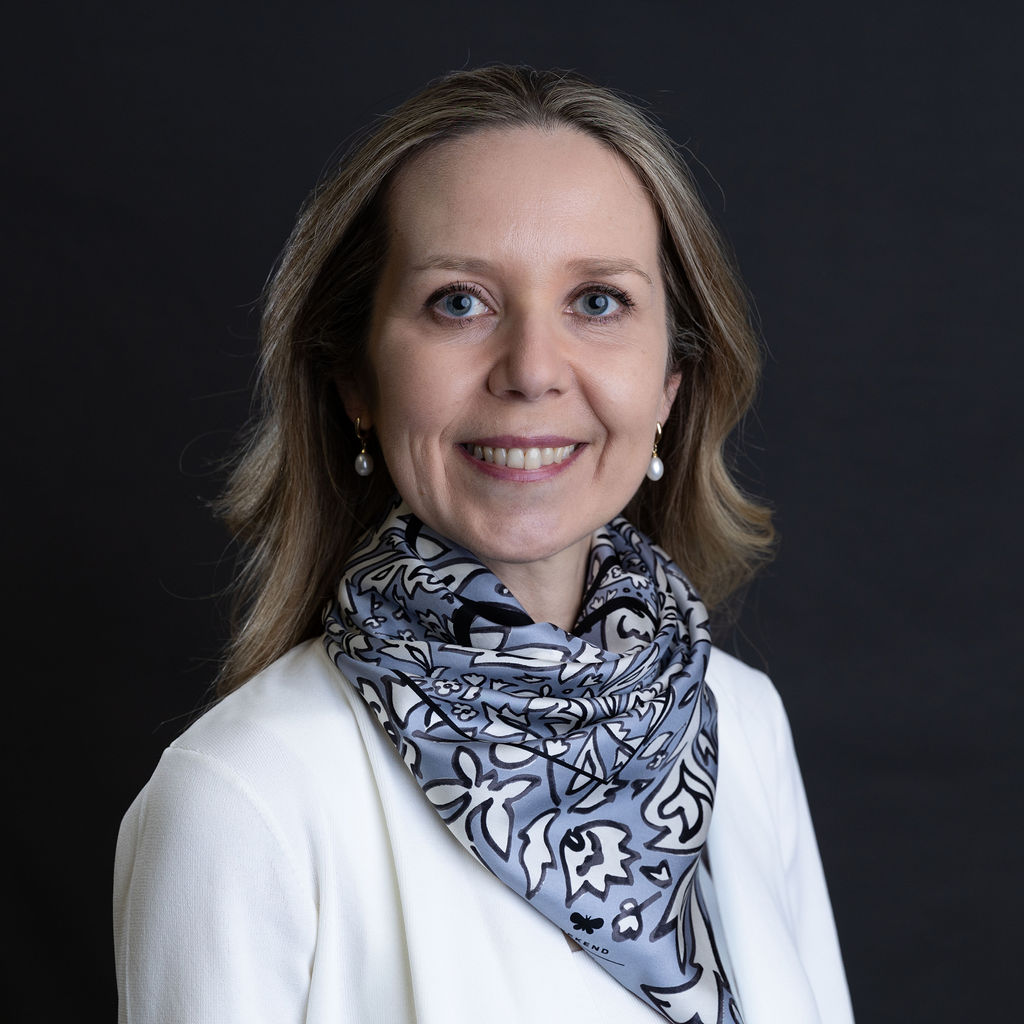 Anastasia Seale, with her long hair and a warm smile, radiates elegance in a white top paired with a patterned scarf. The dark background enhances this professional portrait setting, capturing her poised presence beautifully.