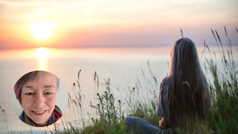Woman sitting in long grass by the sea at sunset, enjoying calm and reflection in nature for improved mental wellbeing