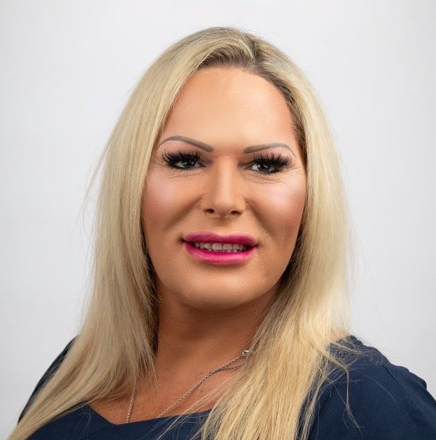A person with long blonde hair is smiling at the camera in our About Us section. They are wearing a dark top and have bright pink lipstick and well-defined eyebrows, set against a plain light background.