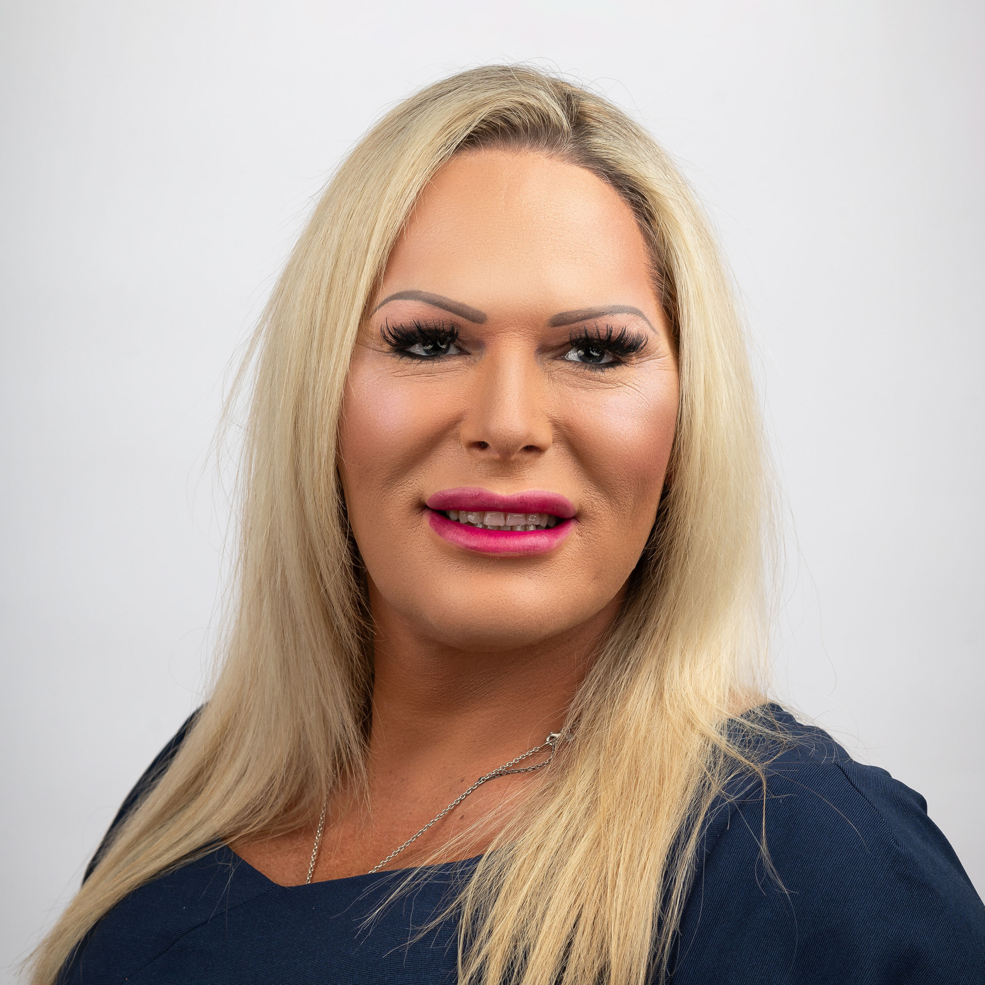 Smiling warmly at the camera, this person with long blonde hair dons a navy blue top against a plain white background—a picture-perfect representation for our About Us section.