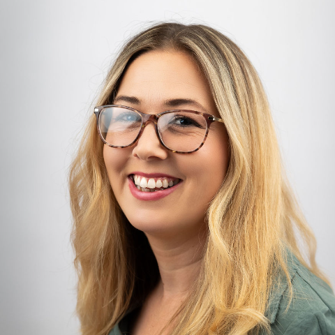 A woman with long blonde hair and glasses smiles at the camera, embodying the friendly spirit of our About Us page. She is wearing a green top, standing against a plain white background.