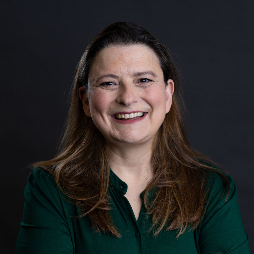 Emma Casey, a woman with long brown hair, smiles warmly in front of a plain dark background. She is wearing a dark green blouse, her arms crossed in a relaxed, confident pose that perfectly captures her approachable demeanor.