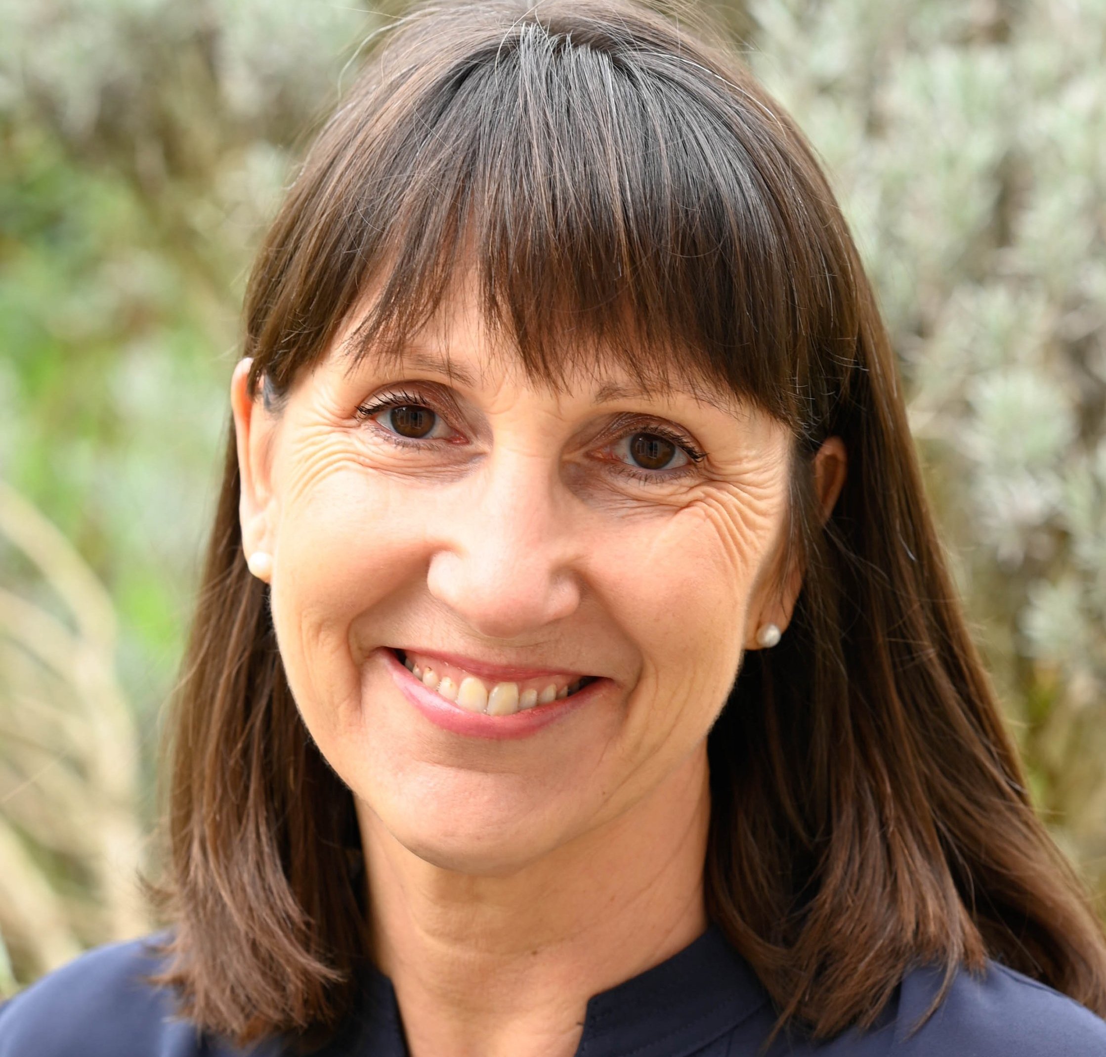 Clare Carter, a woman with straight brown hair and bangs, smiles at the camera. She is wearing a dark top and small stud earrings, with a blurred outdoor background of greenery behind her.