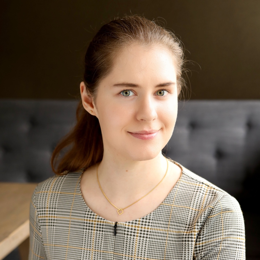 Imogen Wade, with her long brown hair, is smiling at the camera in a plaid top and gold necklace. Shes seated against a softly blurred backdrop that reveals a dark wall and part of a couch.