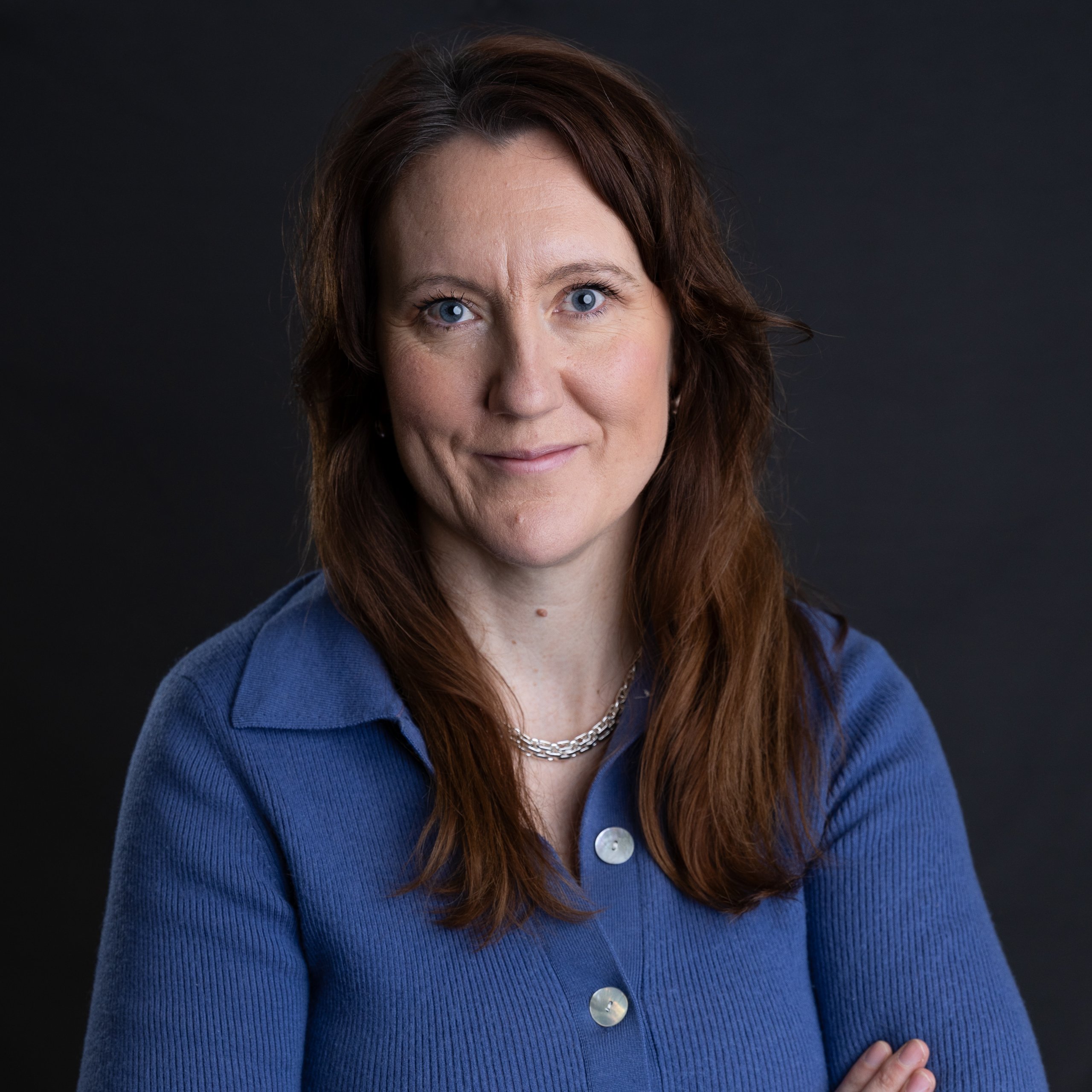 Dr. Charlotte Vaughan, with long brown hair, stands confidently against a dark background. She wears a blue button-up shirt and a silver necklace, arms crossed and slightly.