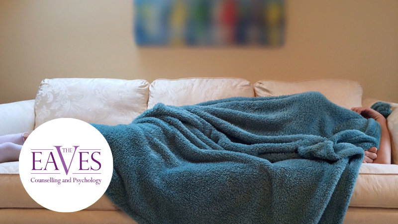 Person lying under a blue blanket on a sofa, illustrating how exhaustion, burnout, or mental health challenges can be mistaken for laziness.