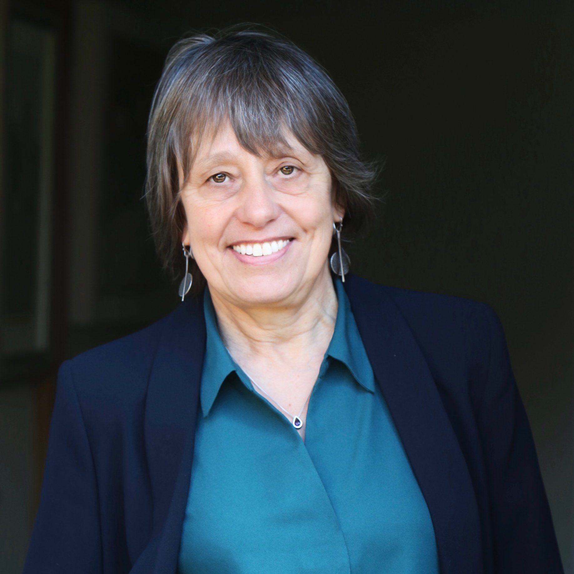 Linda Hoinville, with a radiant smile, stands confidently in a teal blouse and dark blazer against a dim backdrop.