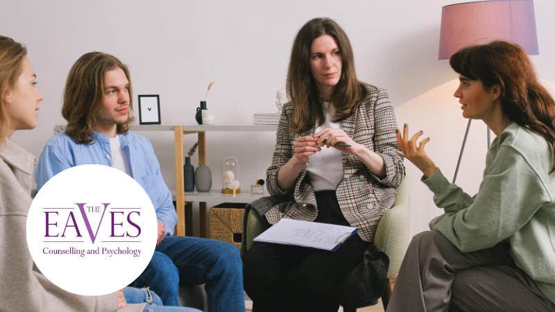 Four adults seated in a circle having a facilitated mental health discussion, with one woman holding a clipboard for notes.