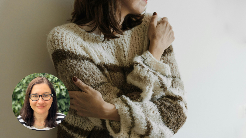 Woman wearing a cream and brown sweater hugging herself and smiling, representing self-compassion, self-care and emotional wellbeing.