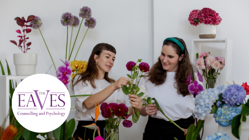 Two young women working together in a flower boutique, arranging pink flowers into a vase, representing creativity, collaboration and neurodiversity at work.