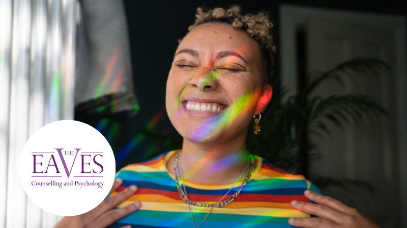 Person smiling with eyes closed, wearing a rainbow-striped shirt. Colorful light reflections create a rainbow effect on their face. To the left is a logo with the text The Eaves Counselling and Psychology. A plant is visible in the background.