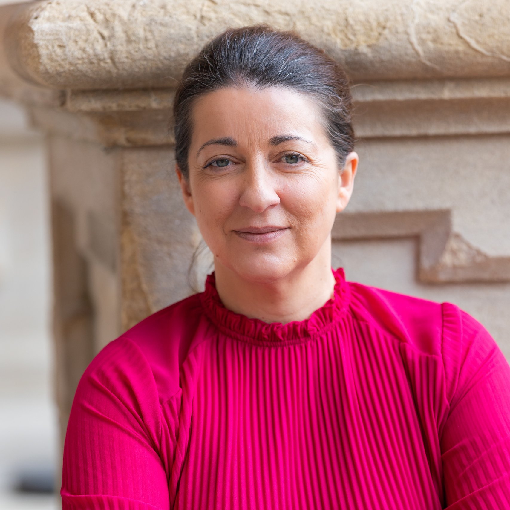 Nadia DiLuzio, with dark hair in a bun, sits against a stone wall wearing a bright pink, pleated blouse. She gazes at the camera with a neutral expression, her hands resting calmly on her lap.