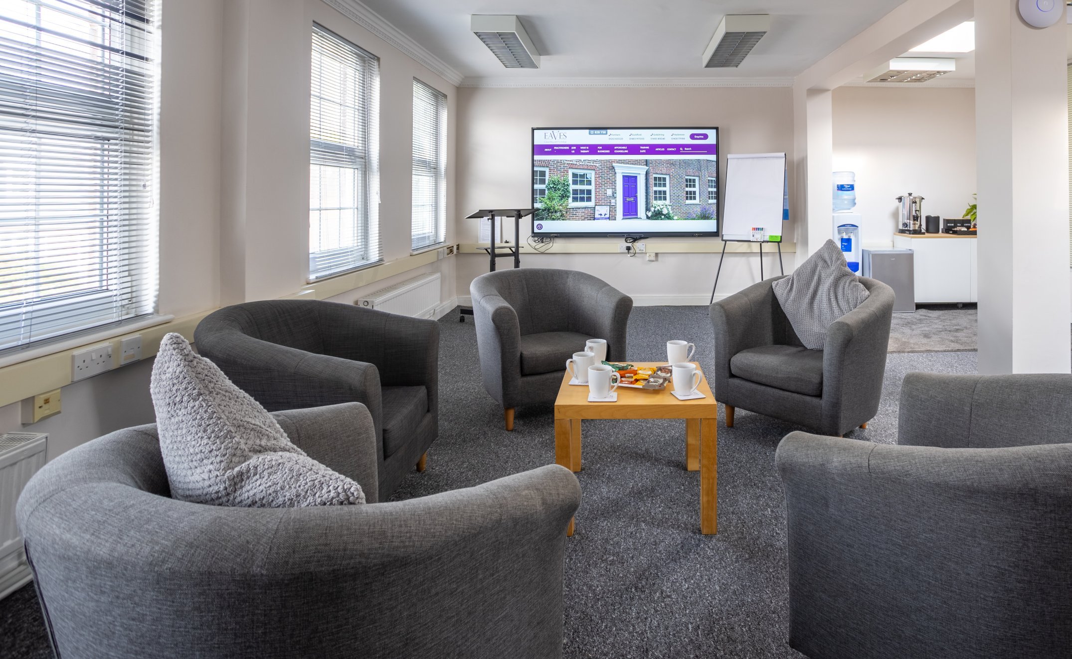 A modern office lounge with four gray armchairs around a wooden coffee table. The table has a coffee pot, cups, and snacks. A screen displays visuals on the far wall. Large windows let in natural light.