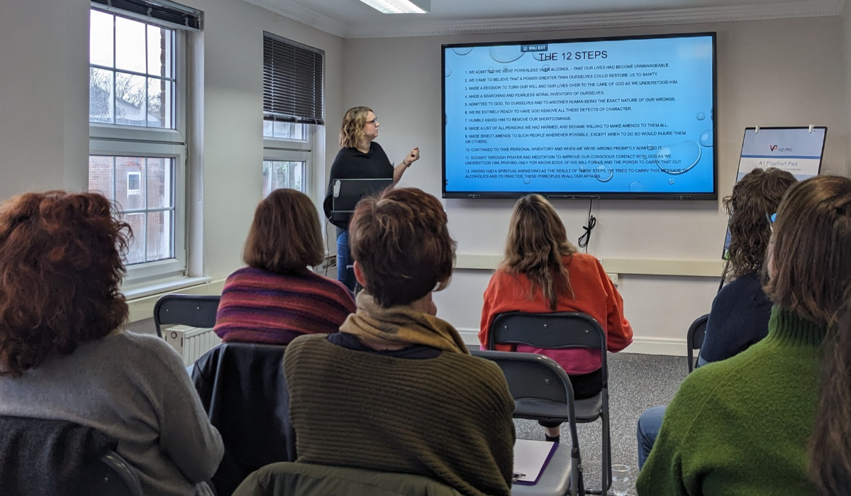 A woman stands in front of a seated audience, pointing at a large screen displaying THE 12 STEPS in bold text. The room has several windows and an easel with a paper pad to the right.