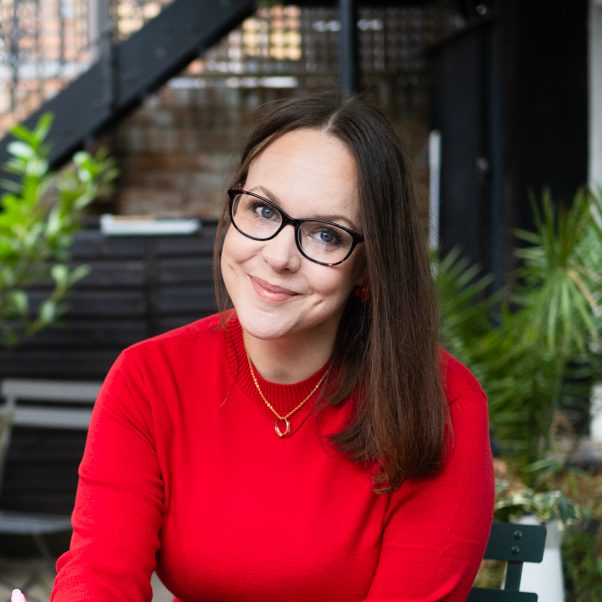 Therapist wearing a red jumper, sitting at an outdoor coffee table surrounded by green potted plants, smiling gently.
