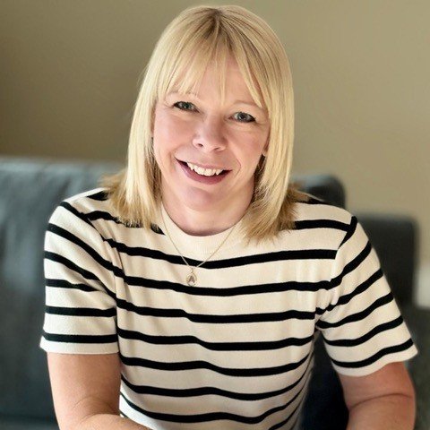 Anna Manners, a woman with shoulder-length blonde hair, is smiling and seated on a gray sofa. She wears a white and black striped shirt, jeans, and a gold bracelet, with her hands clasped in front. The setting features a neutral background and soft lighting.