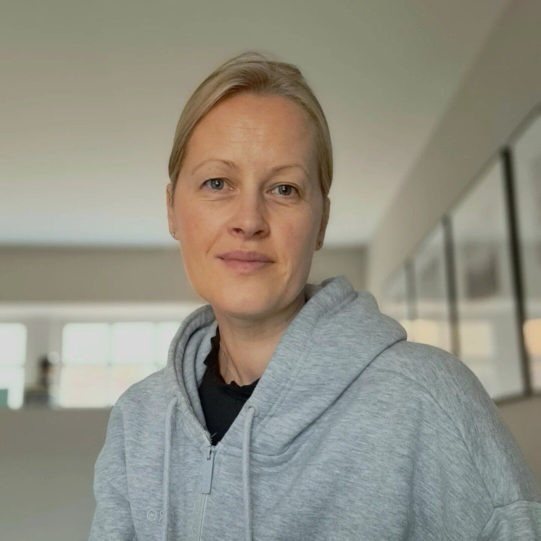 Sarah Sunkin Guildford Counsellor The Eaves. Person with blonde hair wearing a gray hoodie, standing indoors. The background shows blurred framed pictures on a wall and a soft lighting from windows. The person has a neutral expression, looking directly at the camera.