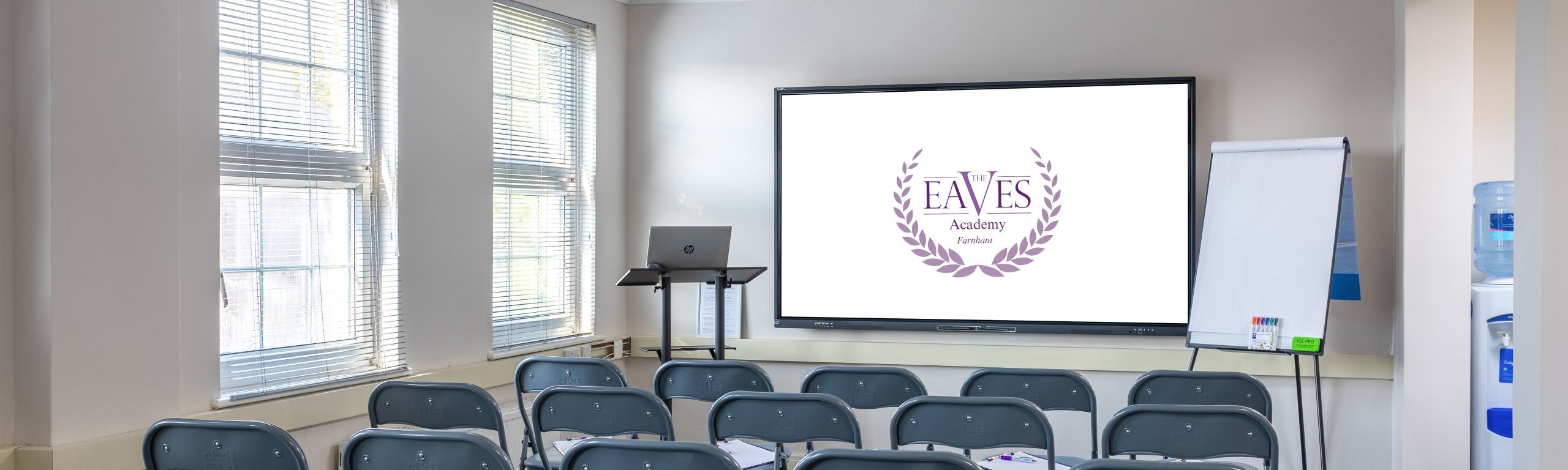 A small conference room with rows of gray folding chairs facing a screen displaying a logo with Eaves Academy and laurel wreaths. A laptop and a flipchart stand on the side, and a water cooler is visible in the corner.