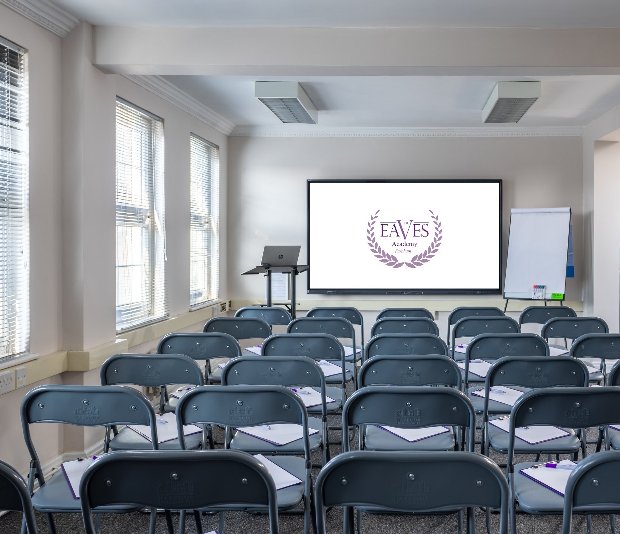 A small classroom setup with rows of folding chairs facing a screen displaying the Eaves logo. Theres a flip chart stand, a laptop on a small table, and a water cooler in the corner. Notepads and pens are placed on each chair.