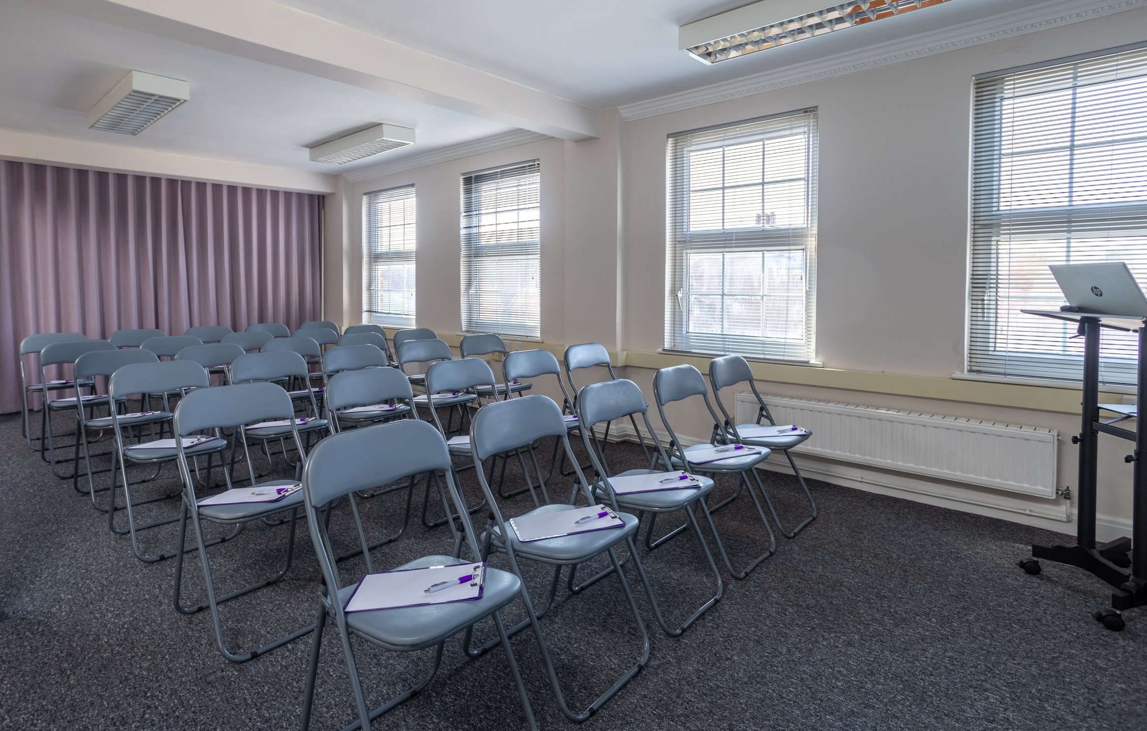 An empty conference room with rows of gray folding chairs, each with a notepad and pen on the seat. Two large windows let in natural light. A small podium with a closed laptop stands near the front.