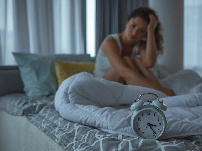 A woman, processing the echoes of trauma, sits on a bed in a dimly lit room, head resting on her hand. An alarm clock displays 5:47 in the foreground while the white comforter and gray patterned sheets add to the quiet weight of her contemplation.