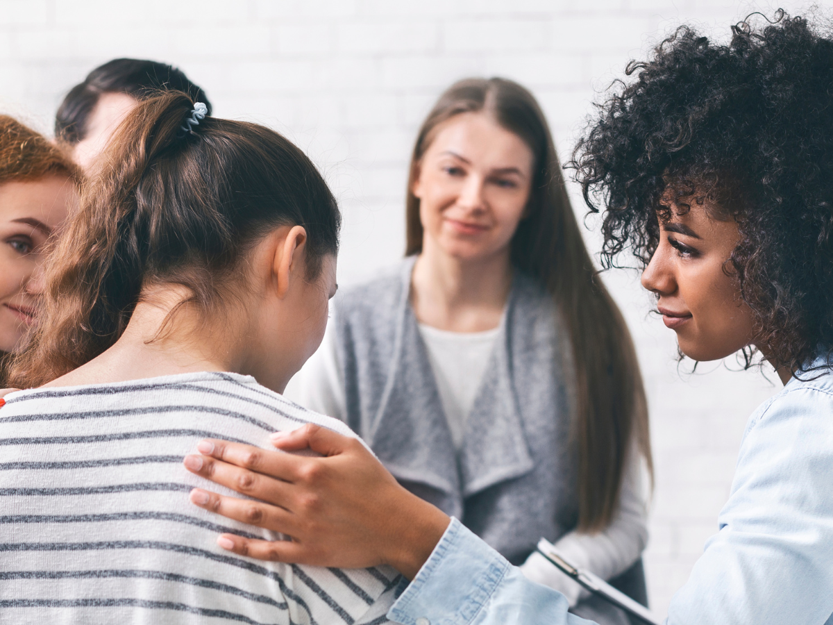 A supportive group meeting where a woman with curly hair gently places her hand on the shoulder of another woman in a striped shirt. Others in the group watch attentively, creating a circle of empathy and understanding in a bright room.