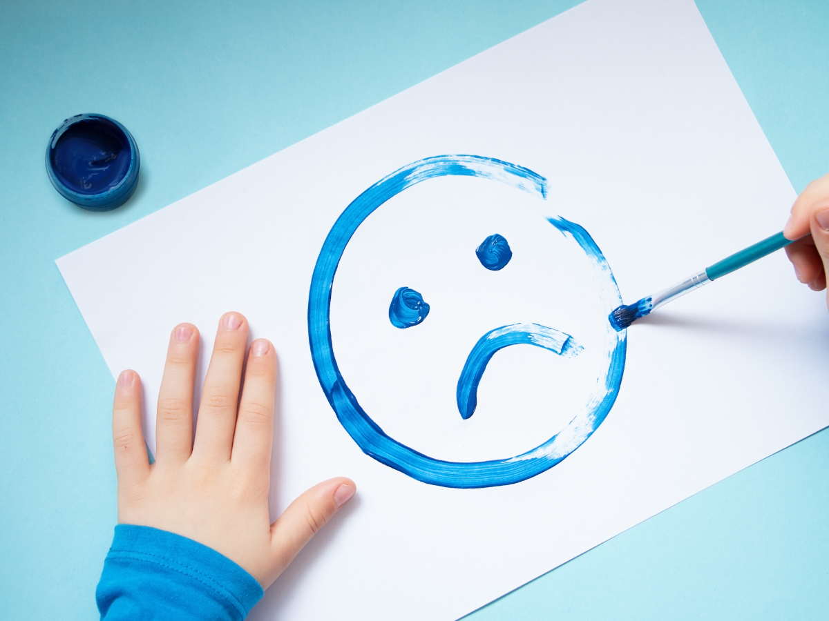 A child’s hands are seen painting a blue sad face on white paper with a paintbrush. An open container of blue paint sits nearby on a light blue surface.