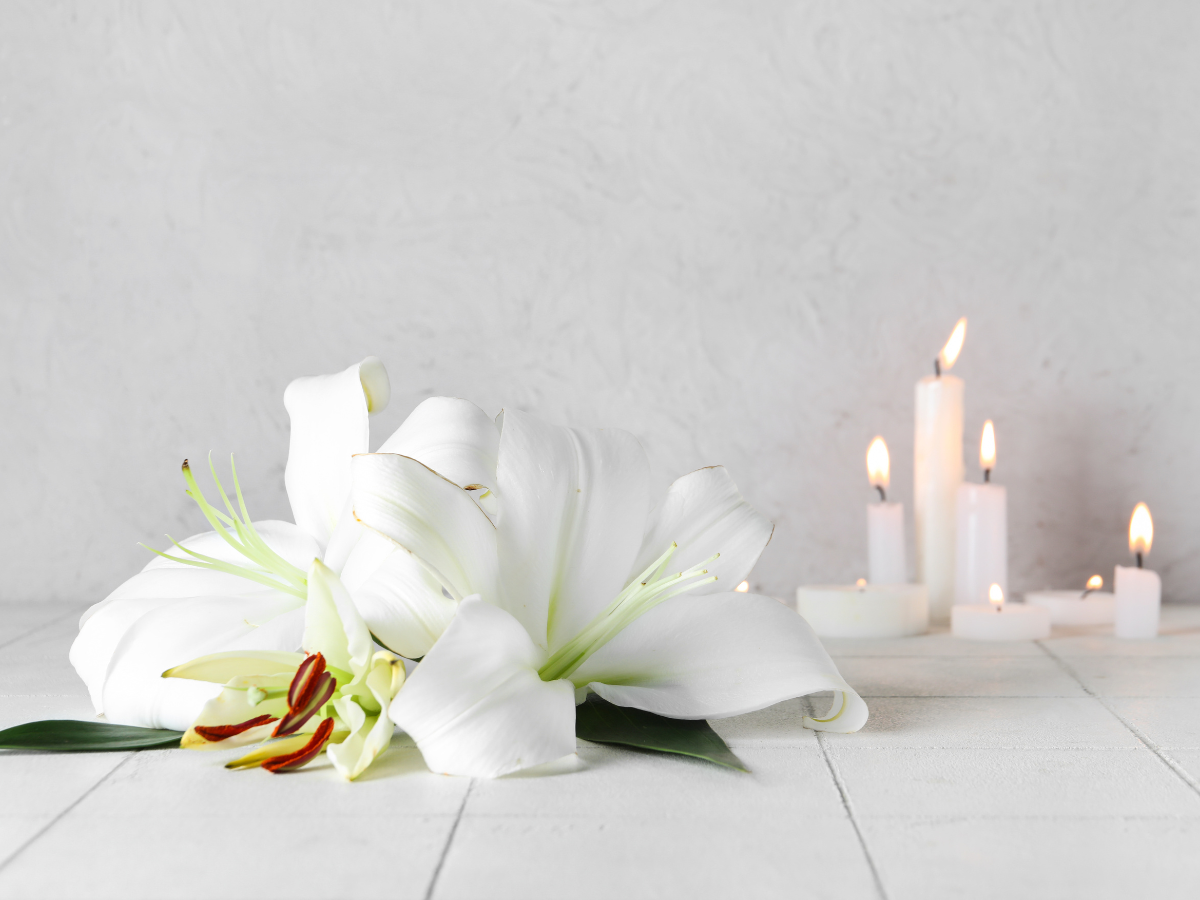 Two white lilies lay on a white surface with green leaves, next to several lit white candles of different heights against a light gray background.