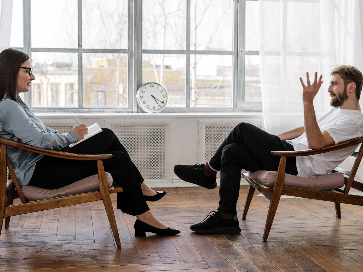 A man and woman sit facing each other in wooden chairs in a bright room. The woman, on the left, takes notes, while the man, on the right, gestures with one hand. A clock on a radiator shelf shows 11:55. Light pours in through large windows.