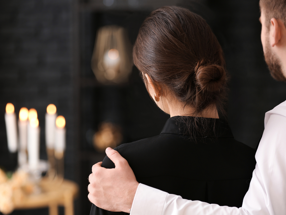 A man gently places his hand on a woman’s shoulder as they stand together, seen from behind. The background is softly lit with candles, creating a somber and comforting atmosphere.