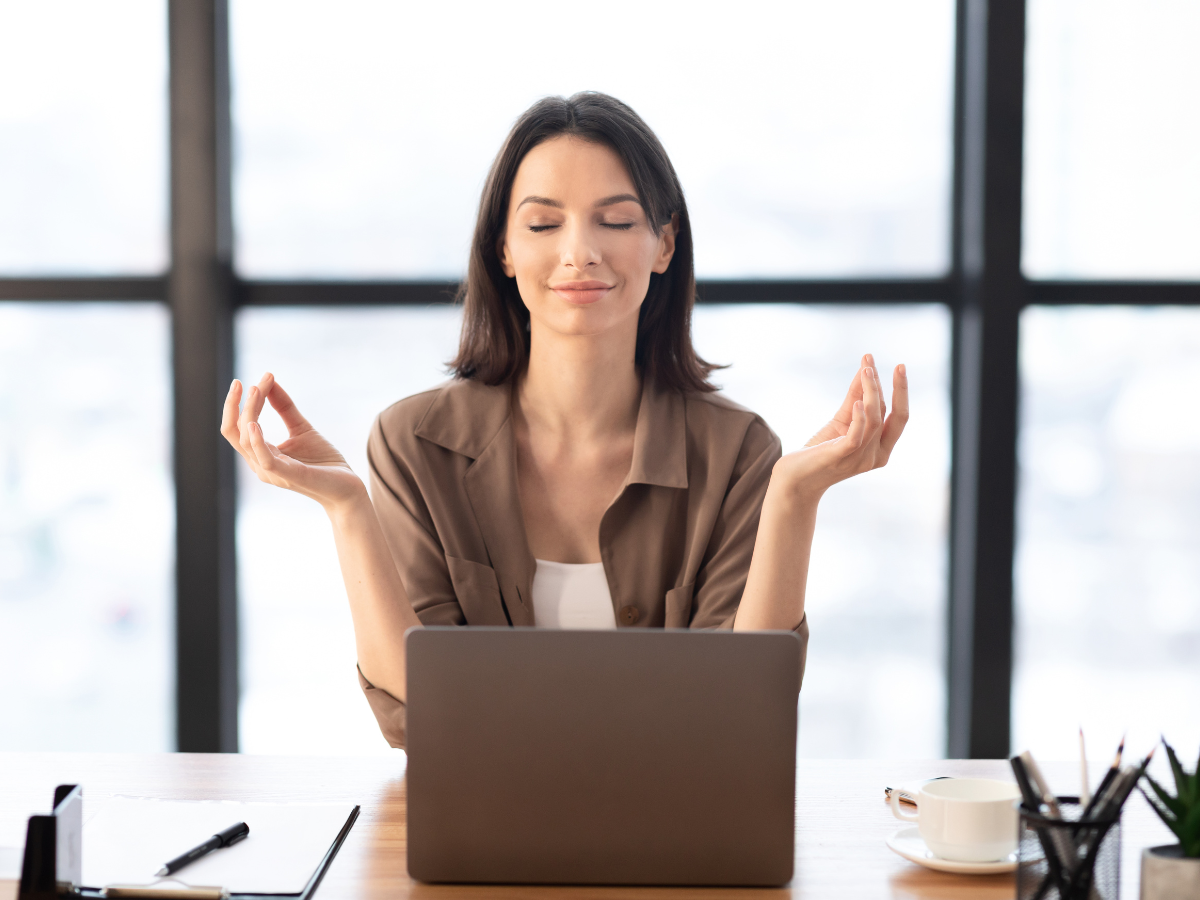 A woman sits at a desk in a meditative pose with her eyes closed, hands raised slightly, and palms facing up. She is in front of a laptop, with a cup and office supplies nearby. Large windows in the background let in bright natural light.
