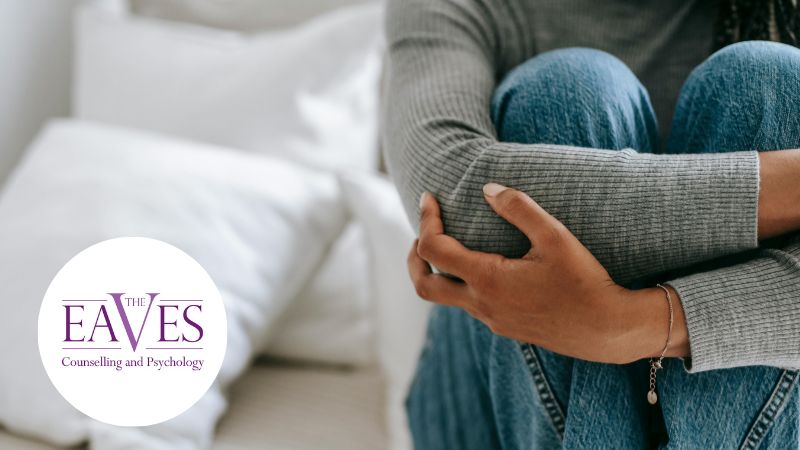 A person in a gray sweater and jeans sits on a light-colored couch, hugging their knees. Their head is not visible. The logo for The Eaves, Counselling and Psychology is on the left side of the image.