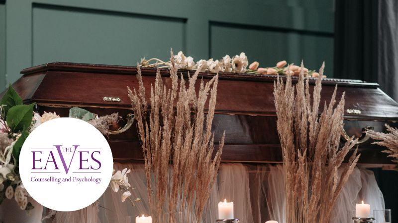 A wooden coffin decorated with flowers and tall dried grass, surrounded by lit candles. The logo for Eaves Counselling and Psychology appears in the lower left corner.