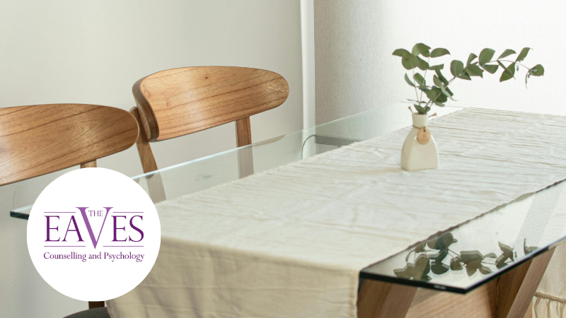 Dining table with wooden chairs and a vase of eucalyptus, symbolising calm and mindful eating environment with The Eaves Counselling and Psychology logo