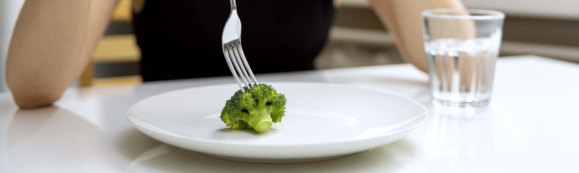 A person sits at a table with a single piece of broccoli on a white plate, holding a fork. Nearby, a glass of water stands ready. The minimalistic meal setup subtly hints at how therapy can help with eating disorders by encouraging mindful eating habits and healthier perspectives on food.