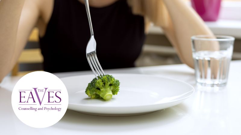 A person sits at a table, contemplating a single piece of broccoli on a large white plate, fork in hand. A glass of water sits nearby. The The Eaves Counselling and Psychology logo suggests how therapy can help with eating disorders by emphasizing mindful eating habits.
