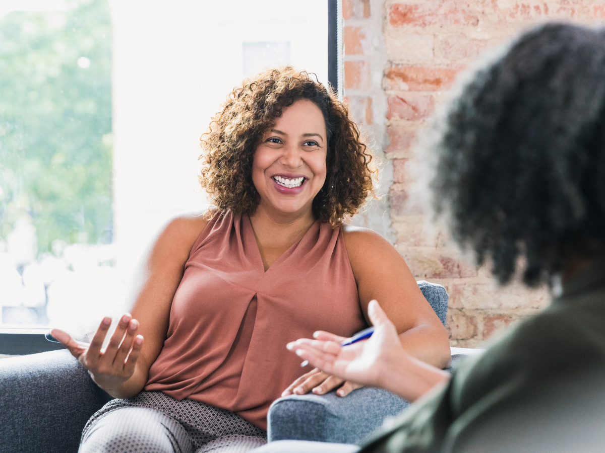 A woman with curly hair in a sleeveless top smiles warmly and gestures as she discusses how therapy can help with eating disorders. They are sitting in a bright room with a brick wall and large windows, creating an inviting atmosphere for open conversation.