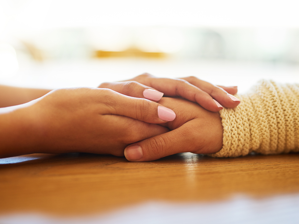 Two people sitting at a table hold hands in a comforting manner, symbolizing the support and connection found in therapy for eating disorders. One person wears a white knitted sleeve, while the other has neatly manicured nails, set against a softly blurred background.